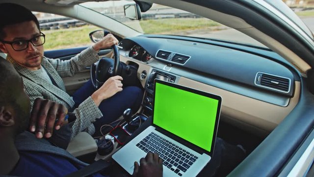African young businessman driving with colleague in car using mock-up greenscreen notebook monitor closeup. Successful multi-race men driving to work together.