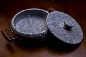 Close up of a smooth round heavy marble stone casserole with brass element and wooden handle with the lid resting half next to it on a brown wooden table surface seen from above looking into the pan