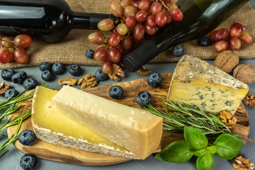 Assortment of cheeses with bottles of wine, herbs, bunch of grapes, nuts and grapes. Food for wine, cheeses delicatessen on a wooden board.
