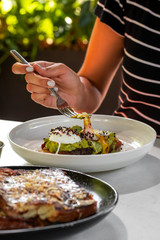 Haute cuisine - grilled sandwich with avocado guacamole sauce and two sunny side up eggs with woman hand and fork outside restaurant in a sunny day