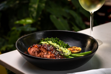 Haute cuisine - grilled salmon with asparagus and cucumbers on a side in a dark bowl outside restaurant in a summer day with a glass of white wine