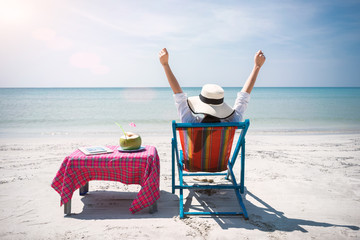 Woman sitting on a cradle of seaside with a clear blue sky. She wore a hat and held her arms up. Her side has coconut water and a laptop.