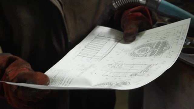 High-class welder is holding detailed drawing of complex device, important part of mechanism to be created, technologically advanced coal boiler is being manufactured, welder is getting ready for work