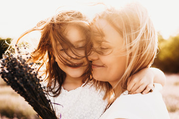 Close up portrait of a lovely little girl embracing her mother smiling outdoor against sunset while wind blowing their hair. - Powered by Adobe