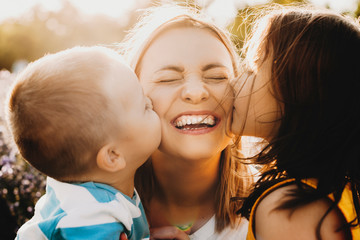 Close up portrait of lovely young mother laughing with closed eyes while her kids is kissing her on...