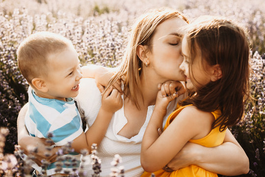 Lovely little girl smiling while her mother is embracing her and kissing while her brother is laughing outdoor in field of flowers.