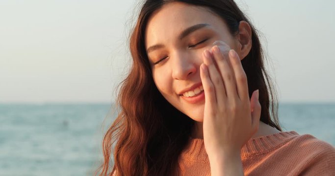 Happy young asian woman is applying a lotion to take care of her skin during a vacation on a beach.