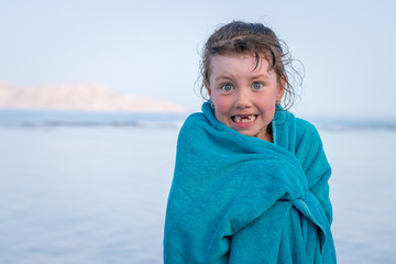 A scared little girl with her front teeth wrapped in a towel is standing on the beach. The child froze after swimming in the sea. Babe basking in a blue towel.