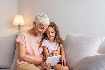 Smiling granddaughter and grandmother with tablet pc computer sitting on couch at home. Picture of cheerful young lady sitting at home with her grandmother using tablet computer.