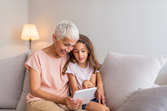 Smiling granddaughter and grandmother with tablet pc computer sitting on couch at home. Picture of cheerful young lady sitting at home with her grandmother using tablet computer. - Powered by Adobe