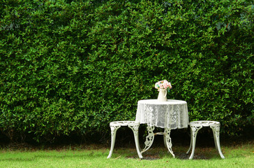 White round table cover with white crochet tablecloth with old pink fabric flowers in white vest on in the garden, green nature background
