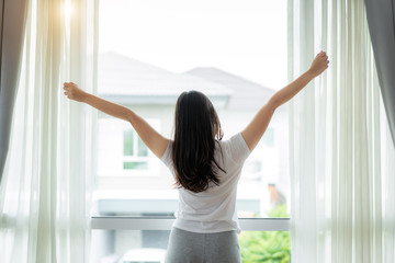 Rear view of Asian Woman stretching hands and body near window after wake up in bedroom at home. Concept for start new day with happiness. Young happy working female life