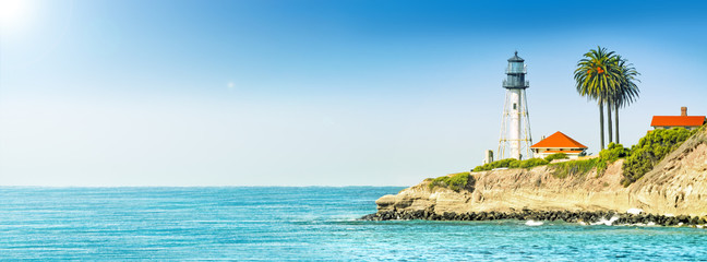 lighthouse tower on cliff in ocean landscape against stormy sea water and blue sky background. Panorama ultra wide side view of marine light beacon with palm tree located on rock. Copy space banner