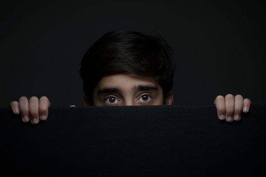 Portrait of a young boy hiding his face behind a black cardboard. (Children) 	