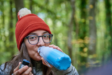 a white Caucasian girl, in the forest drinking water from a blue bottle and looking directly at the camera