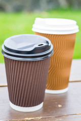 Two paper coffee cups on green background. Two different cups stand on a wooden bench. Green grass background. Spring beautiful day. Tolerance to appearance. Nice look, one thing. Wine coffee. 