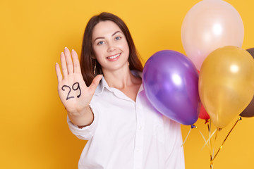 Young woman with multi colored ballons, being excited, wearing white elegant blouse, posing isolated over white background, showing palm with inscription twentie nine, celebrating her birthday. © sementsova321
