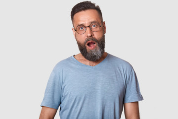 Indoor shot of bearded Caucasian man notices something unexpected, keeps mouth widely opened, being surprised, male posing isolated over white concrete wall. Shock and people emotions concept.