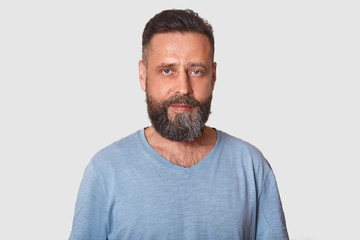 Studio shot of middle aged man with beard dresses grey casual t shirt, posing isolated over light background, attractive male looking directly at camera with calm facial expression. People concept.