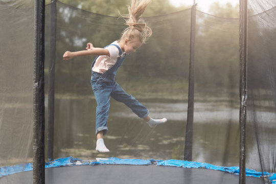 Full length photo of happy girl jumping high on trampoline in park, blonde female chicd with ponytail wearing white shirt and denim overalls, kid spending time in open air, having fun outdoor.