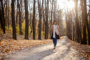 Fototapeta premium Hello autumn. Full length portrait of pensive elegant young woman in coat and scarf with yellow leaves outdoors in the autumn park.