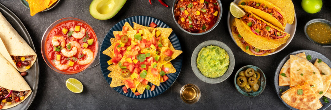 Mexican Food Overhead Panoramic Shot, Many Dishes Of The Cuisine Of Mexico, Flatlay On A Dark Background. Nachos, Guacamole, Shrimp Cocktail, Tacos, Chili Con Carne