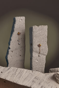 Close-up shot of a pair of stud earrings fixed on the gray half-sawn stones. Each earring is made as a golden bird on the flowers with crystals, jeweller's enamel and a chain with a shimmering pearl. 