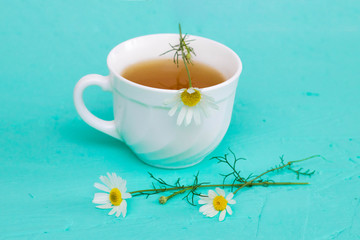 Chamomile tea in a white tea cup, chamomile flowers on the table, turquoise background.