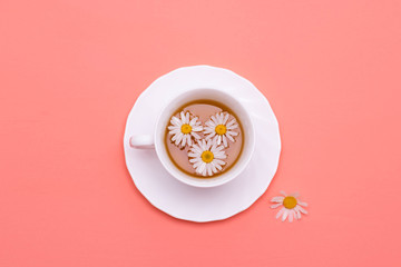 Chamomile tea in a white tea cup, chamomile flower on the table. Pink background, top view.