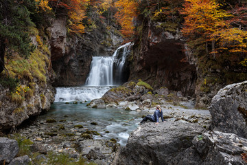 woman with blue jacket watching a waterfall in an autumn mountain forest