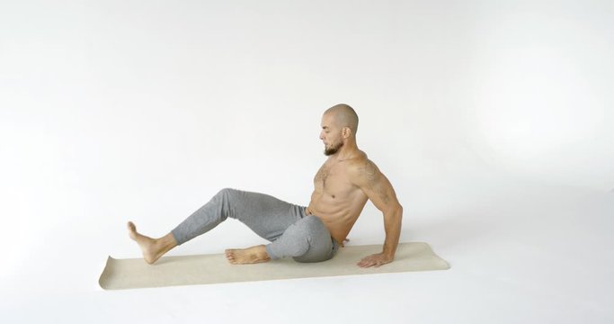 bald man with beard is sitting on floor mat in yoga class and stretching legs muscles, holding foot
