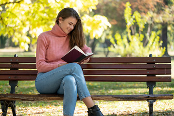 Obraz premium Young woman sitting on a bench in autumn park and is reading a book.