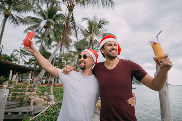 Young gay couple with cocktails in Christmas caps on the ocean on a tropical island.
