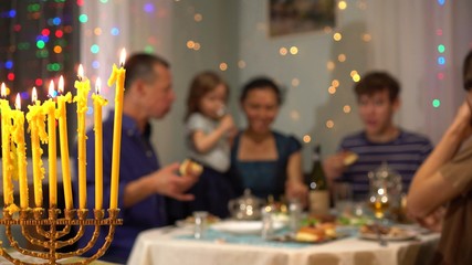 Happy Jewish family celebrates Hanukkah holiday. Hanukkah lights at home, with family and friends. Chanukah table dinner and foods. Menorah with nine burning candles in the foreground