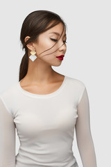 Naklejka premium Medium shot of a young Asian woman in an ivory white blouse and with a golden stud earring made as golden and nacre rhombus-shaped plates.