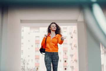 Smiling positive caucasian woman in sportswear and with curly hair carrying bag and going to gym. City life.