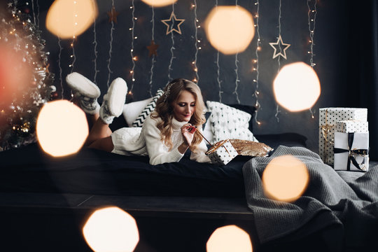 Stock photo of beautiful blonde woman in white knitted dress and slippers relaxing on the bed with pillow. The bed is decorated with cushions. The wall is decorated with garland. Christmas tree in