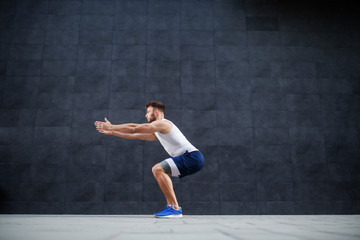 Side view of handsome muscular caucasian man in shorts and t-shirt doing squatting exercise...