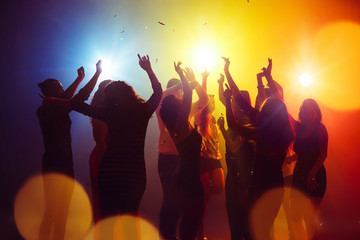Confetti. A crowd of people in silhouette raises their hands on dancefloor on neon light...