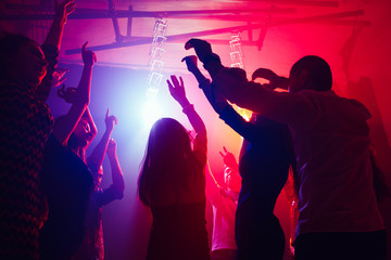 Cheers. A crowd of people in silhouette raises their hands on dancefloor on neon light background. Night life, club, music, dance, motion, youth. Purple-pink colors and moving girls and boys.