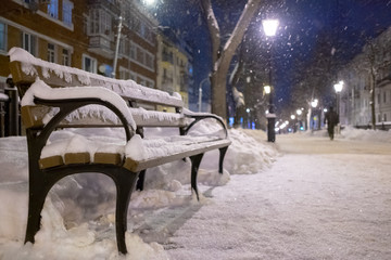 Fototapeta na wymiar A bench covered with snow on a night boulevard in the city in the light of lanterns. Winter city landscape.
