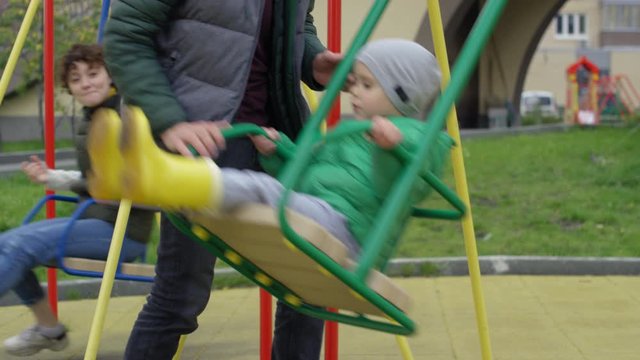 Full shot of happy young Caucasian child in warm jacket and boots riding on swing in playground, unrecognizable father standing by and rocking him, and smiling mom in background