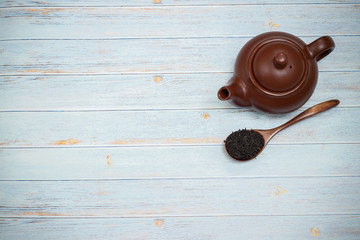 Brown clay teapot on a blue wooden background. Nearby are tea leaves on a wooden spoon.