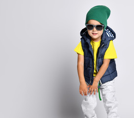 Stylish cheerful little boy 4 years old in sweatpants, a green hat, glasses and a down vest has fun
