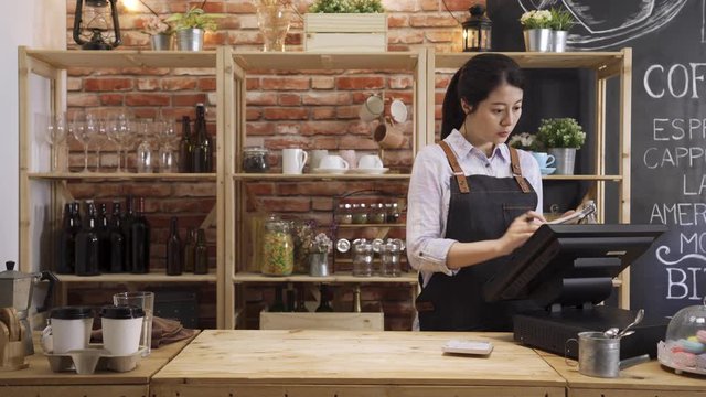 confident asian chinese woman bartender taking note and counting on calculator in cafe counter bar table. elegant beautiful female barista using point of sale terminal machine in coffee shop