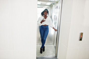 Fototapeta na wymiar Stylish african american women in white blouse and blue jeans posed at elevator with mobile phone at hand.