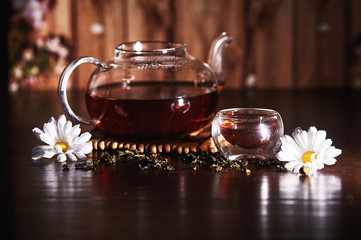 Glass teapot with tea and daisies on dark wooden background