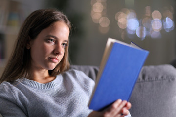 Serious woman reading a paper book on a couch in the night