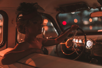 Portrait of beautiful sexy fashion blond girl. Model with bright makeup and curly hairstyle in retro style hat sitting in old car. Attractive woman  rides around the night city