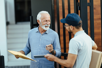 Happy mature man talking to courier while signing on touchpad for a delivery.